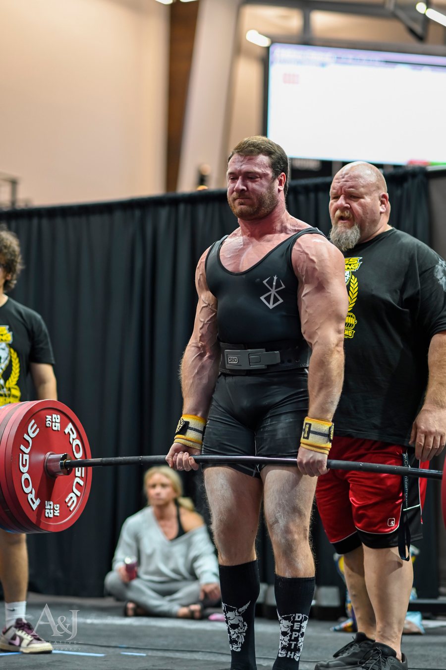 Dr. Addison DeBord locking out a deadlift at a powerlifting meet while a spotter looks on.